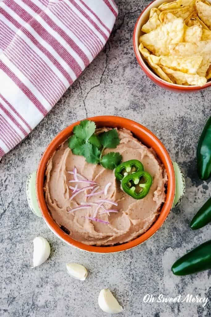 overhead shot of bowl of party bean dip topped with cilantro, jalapeno slices, and red onion