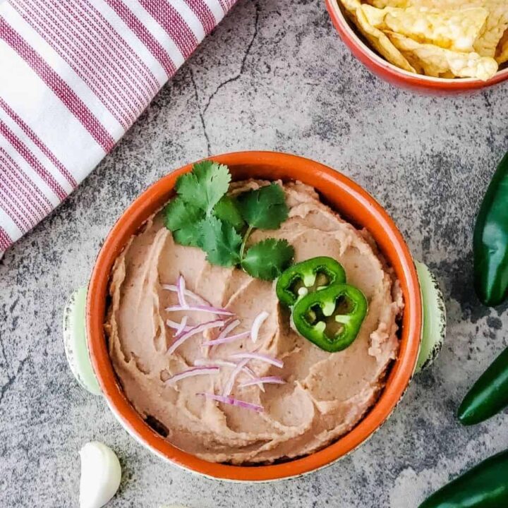 overhead shot of bowl of party bean dip topped with cilantro, jalapeno slices, and red onion