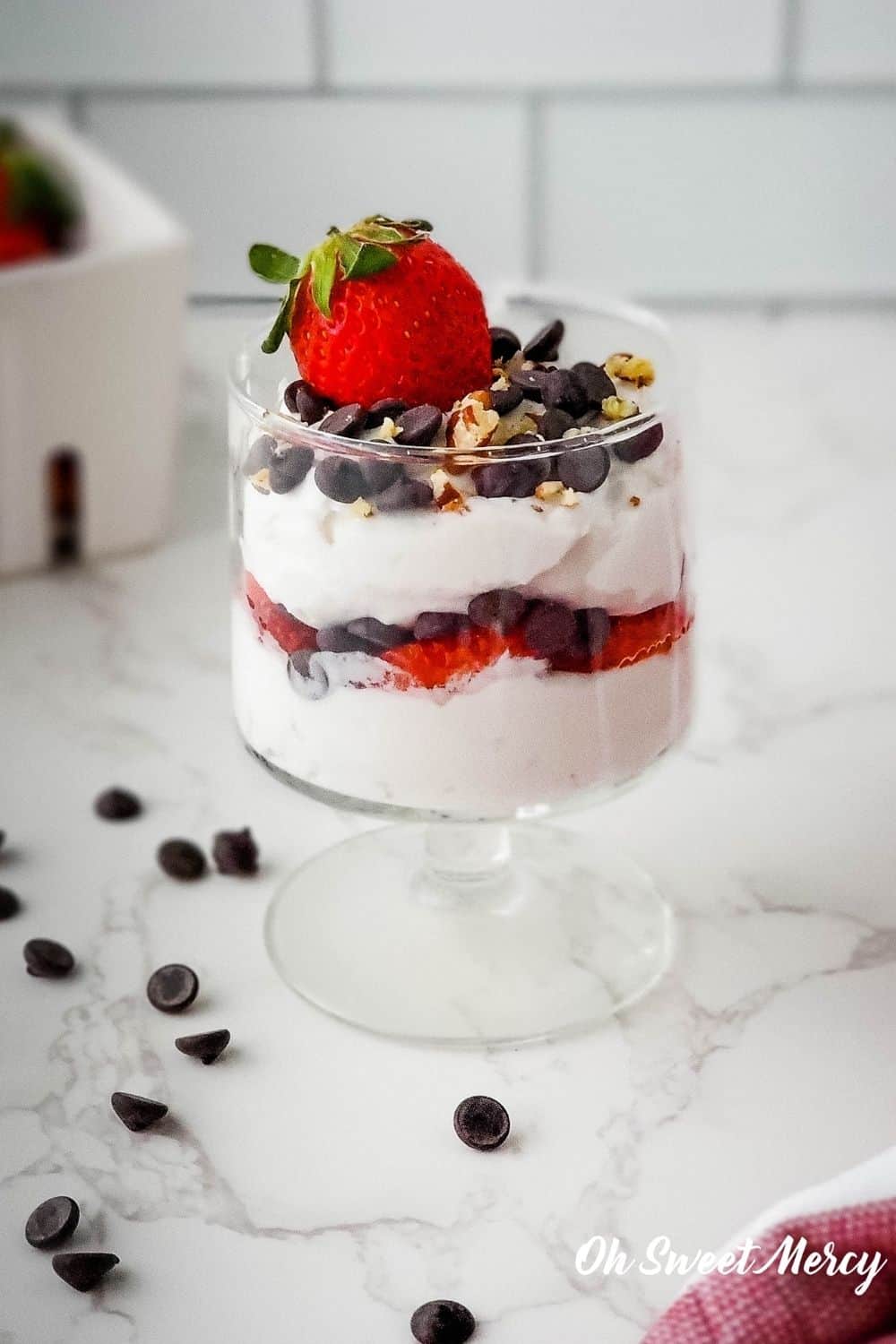 Parfait glass of Special Occasion Chocolate Chip Parfait showing layers of fluffy cream cheese, strawberries, chocolate chips, topped with chocolate chips, a whole strawberry, and sprinkle of pecan pieces