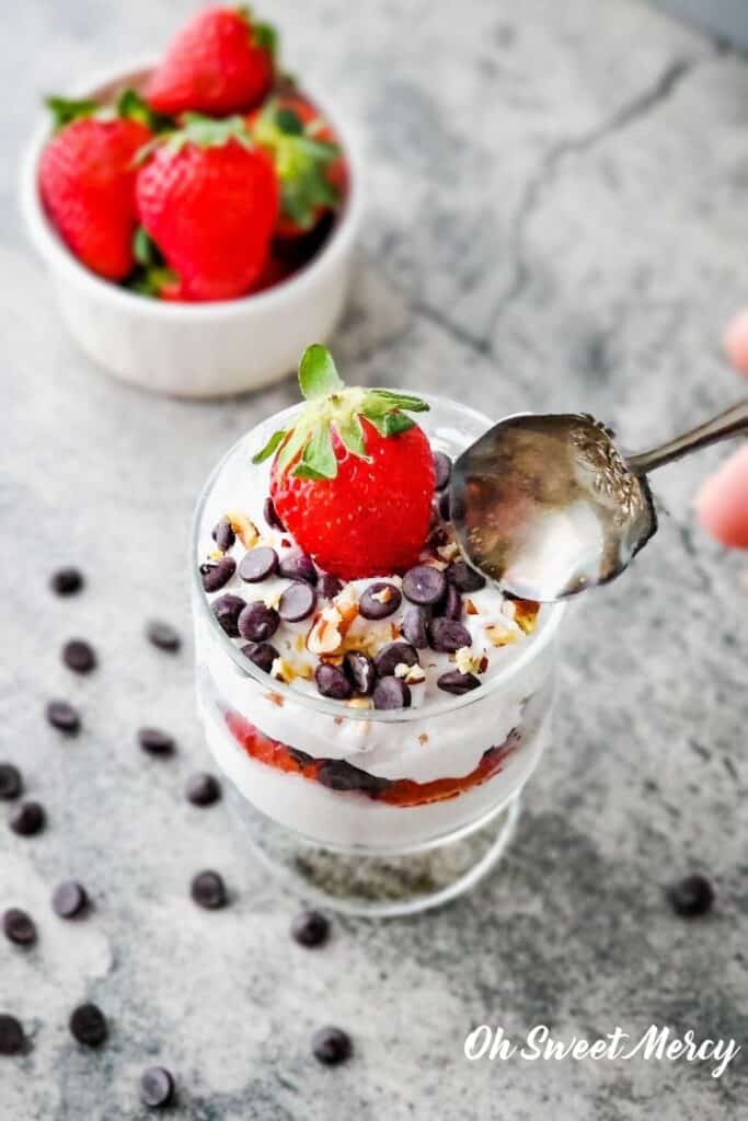 Special Occasion Chocolate Chip Parfait topped with whole strawberry, chocolate chips, pecan pieces, with a decorative vintage spoon about to scoop into it. Bowl of strawberries in the background, chocolate chips scattered around.