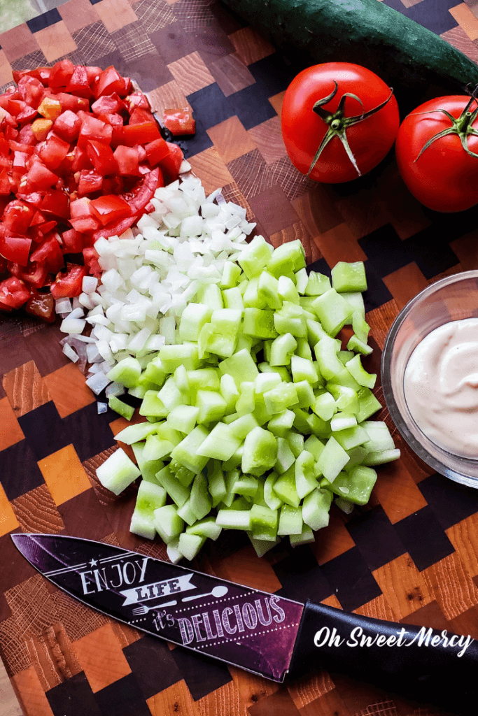 Creamy Cucumber Tomato Salad ingredients on chopping board. Diced tomatoes, onions, cucumbers, bowl of dressing, knife.