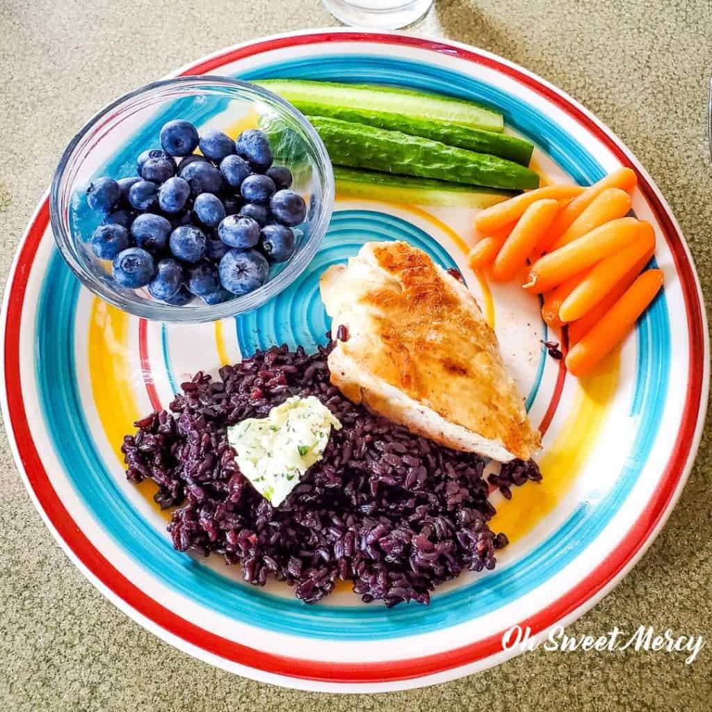 Colorful plate of grilled chicken, fresh cucumbers, carrots, cooked black rice, pat of herbed butter and fresh blueberries