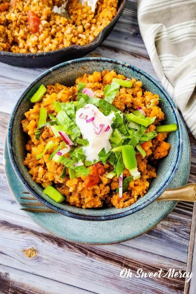 Bowl of low carb taco skillet made with cauli rice, topped with lettuce, sour cream, and onions