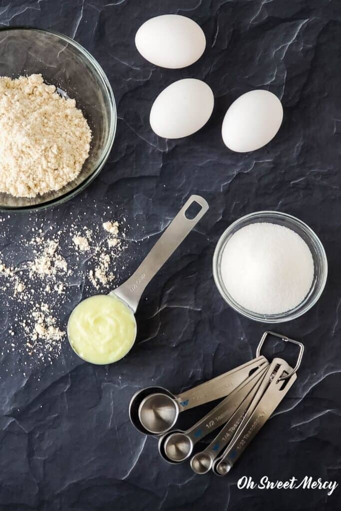 Overhead shot of buttery coconut oil surrounded by bowl of flour, eggs, bowl of sweetener, and set of measuring spoons