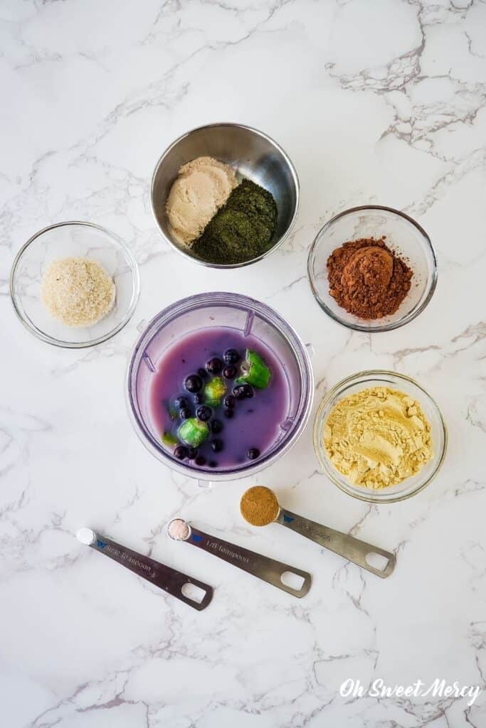 Overhead shot of blender cup with nut milk, blueberries, and okra, surrounded by psyllium husk, baobab, Dynamic Duo Greens Powder, cacao powder, vegan protein powder, camu camu powder, salt, and stevia.