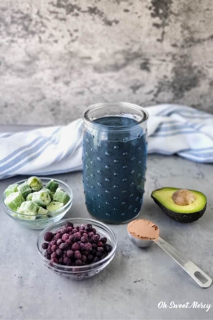 Glass of my Chocolate Blueberry Smoothie (AKA The Breakfast Smoothie) with some ingredients that are in it: blueberries, okra, cacao powder, avocado