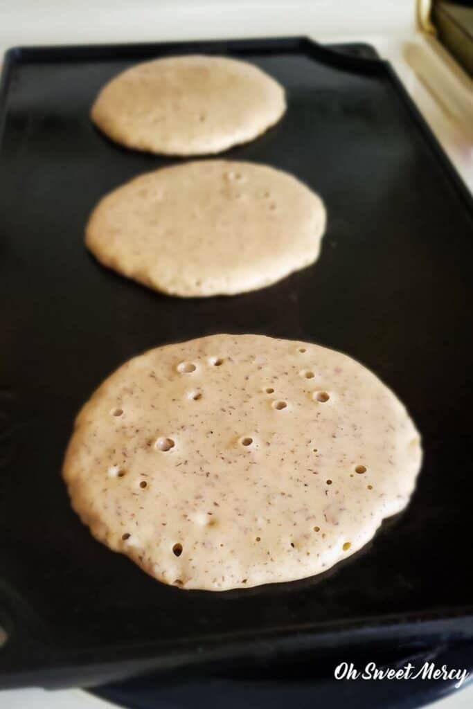 3 millet pancakes cooking on a cast iron griddle, not flipped, with bubbles in batter