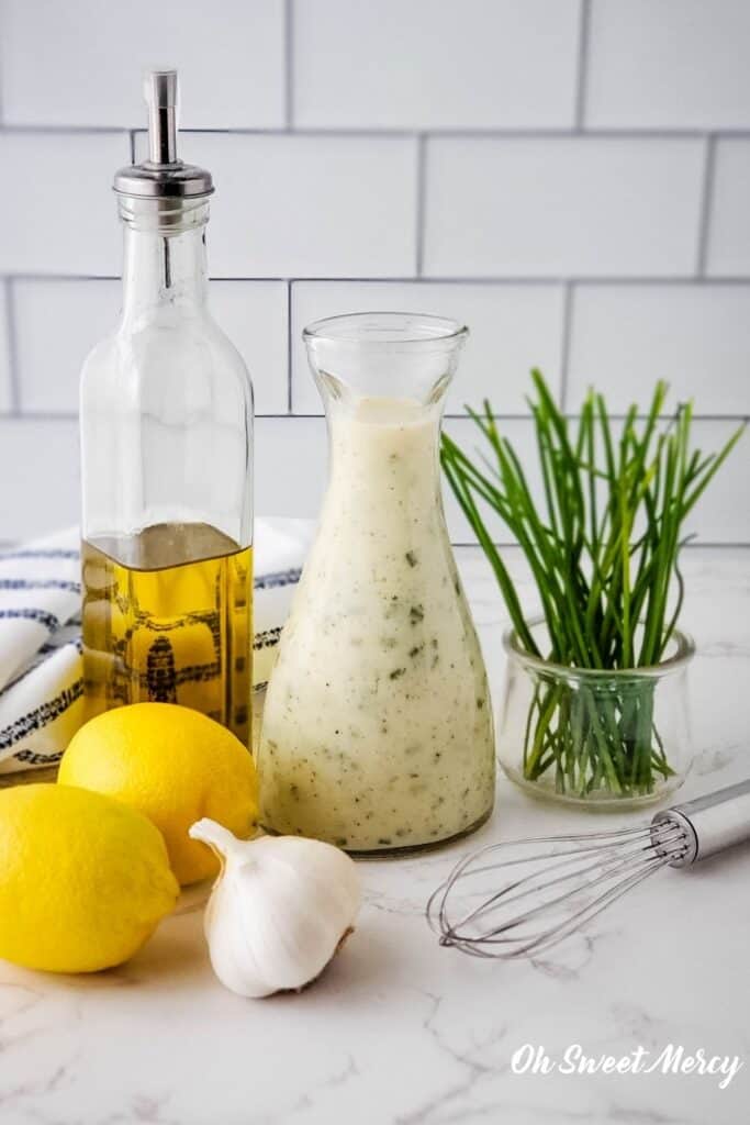 Bottle of Creamy Lemon Garlic Salad Dressing with olive oil, lemons, garlic, chives, and whisk on kitchen counter