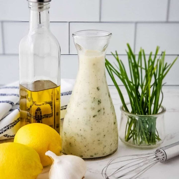 Bottle of Creamy Lemon Garlic Salad Dressing with olive oil, lemons, garlic, chives, and whisk on kitchen counter