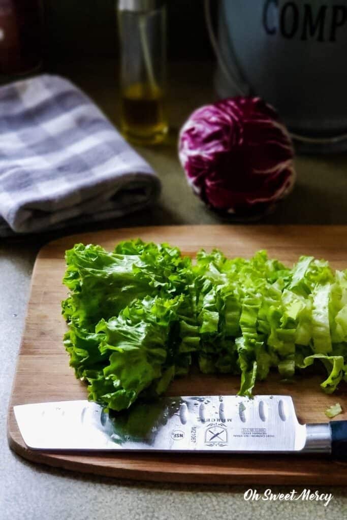 Lettuce on cutting board with knife, towel, bottle of olive oil, head of radiccio in background