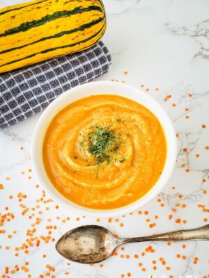 Overhead shot of white bowl with Red Lentil and Delicata Squash Soup (a blended soup) in a white bowl on white marble background, a spoon, dry red lentils scattered around, a delicata squash on top of a folded towel.