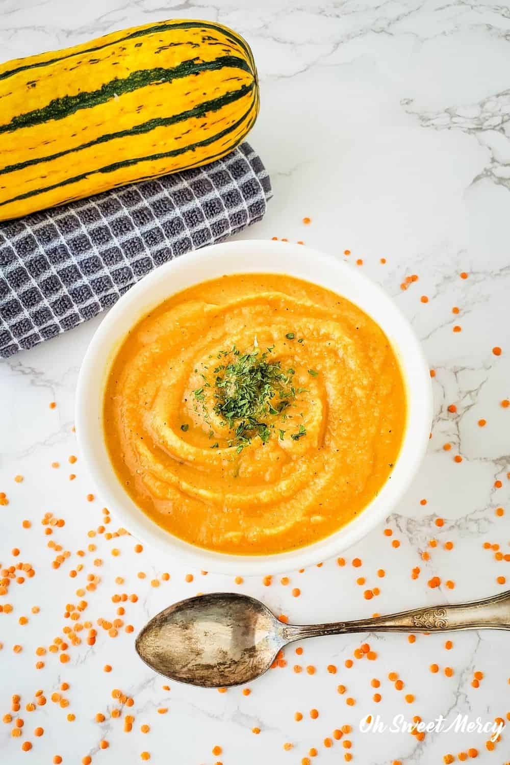 Overhead shot of white bowl with Red Lentil and Delicata Squash Soup (a blended soup) in a white bowl on white marble background, a spoon, dry red lentils scattered around, a delicata squash on top of a folded towel.