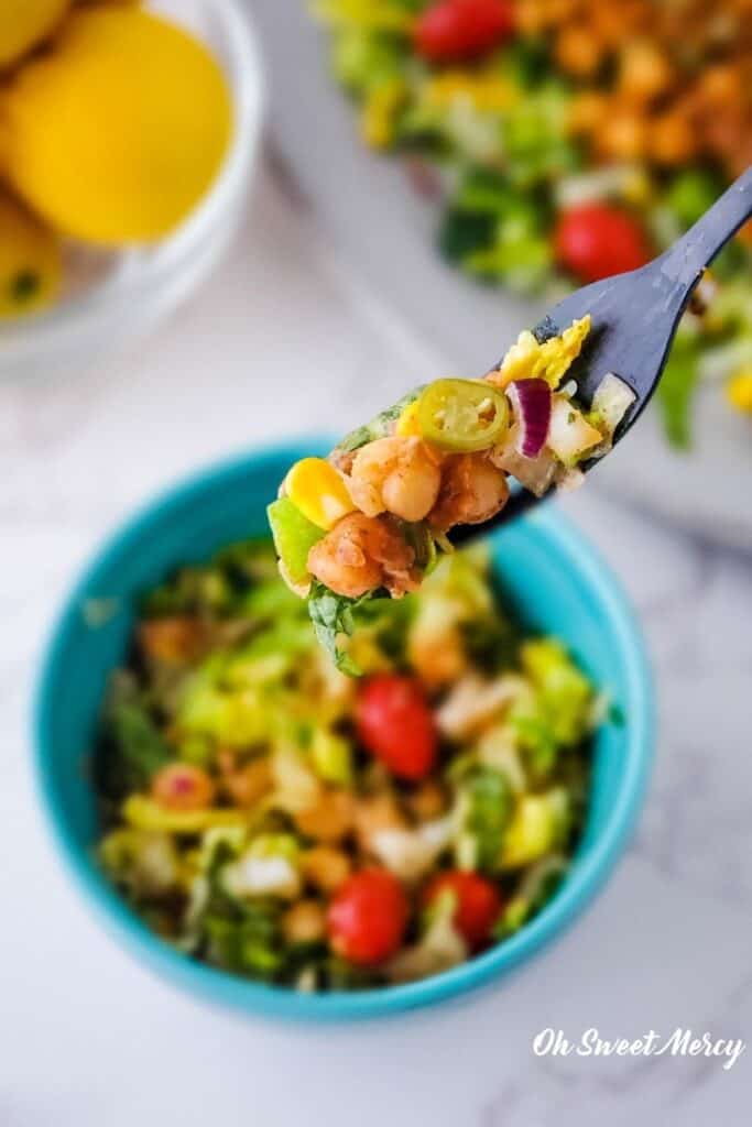 Low Fat Chickpea Taco Salad on fork with teal bowl of salad, bowl of lemons, and platter of salad in background.