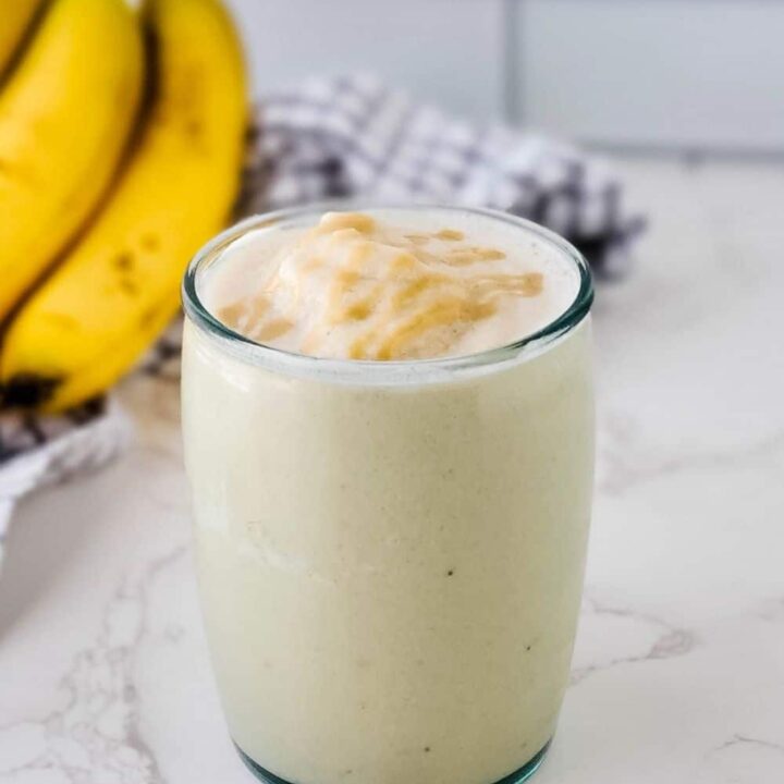 Glass of Banana Maple Tahini Smoothie on kitchen counter with bunch of bananas in background