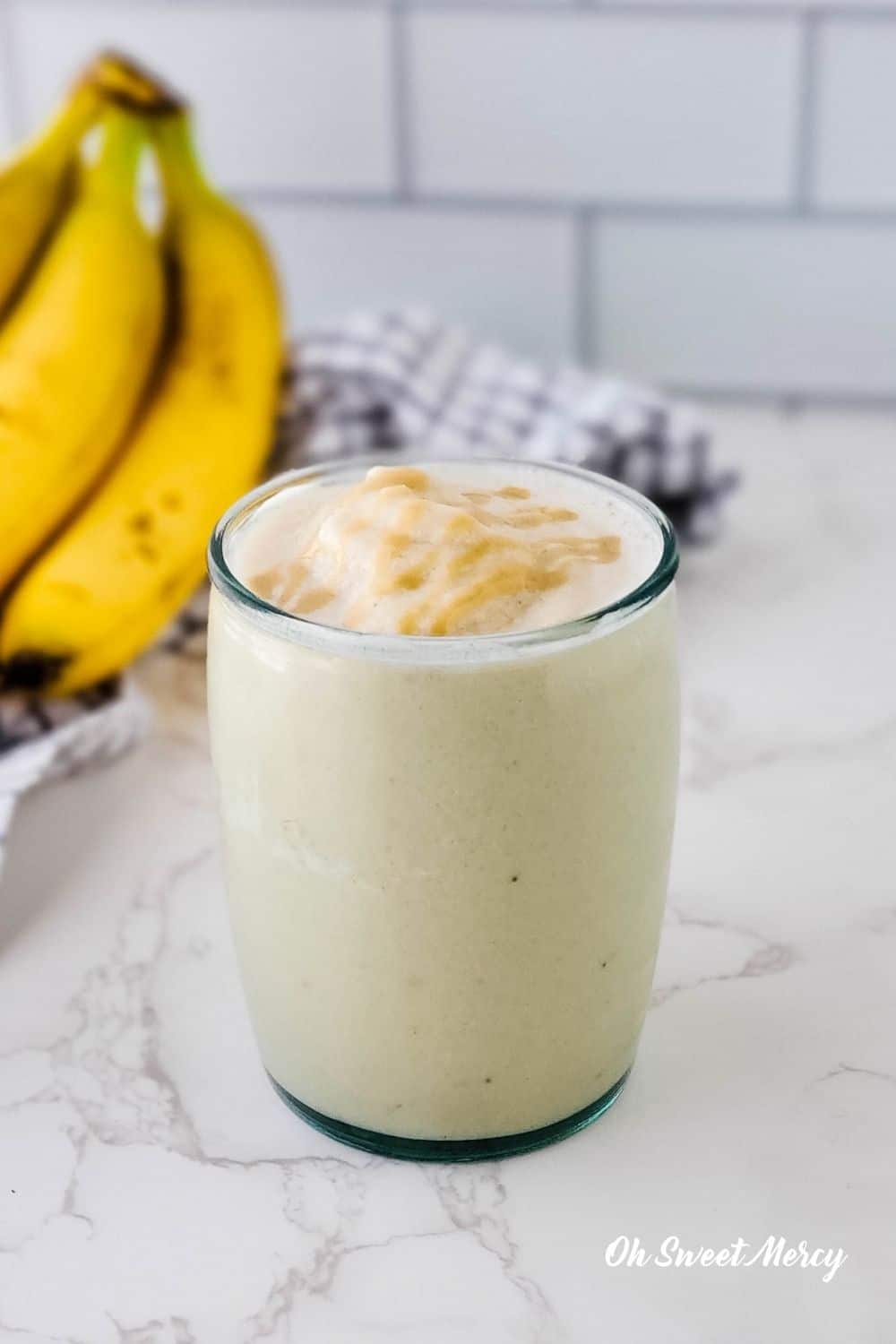 Glass of Banana Maple Tahini Smoothie on kitchen counter with bunch of bananas in background