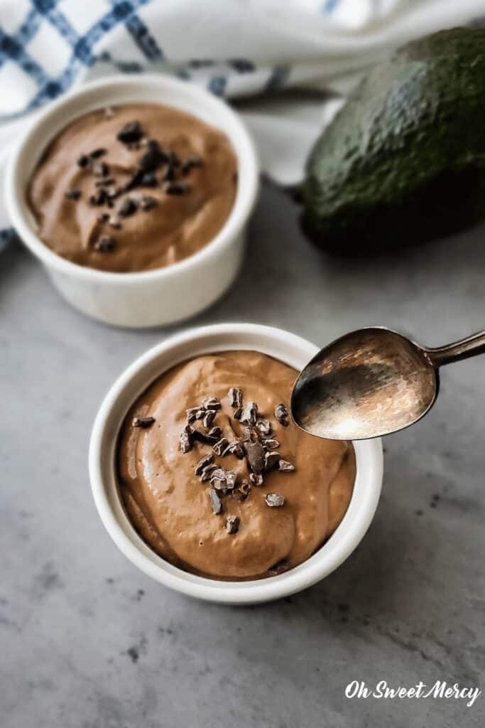 Small white bowls of pudding with raw cacao nibs sprinkled on top, a spoon hovering over one of the bowls
