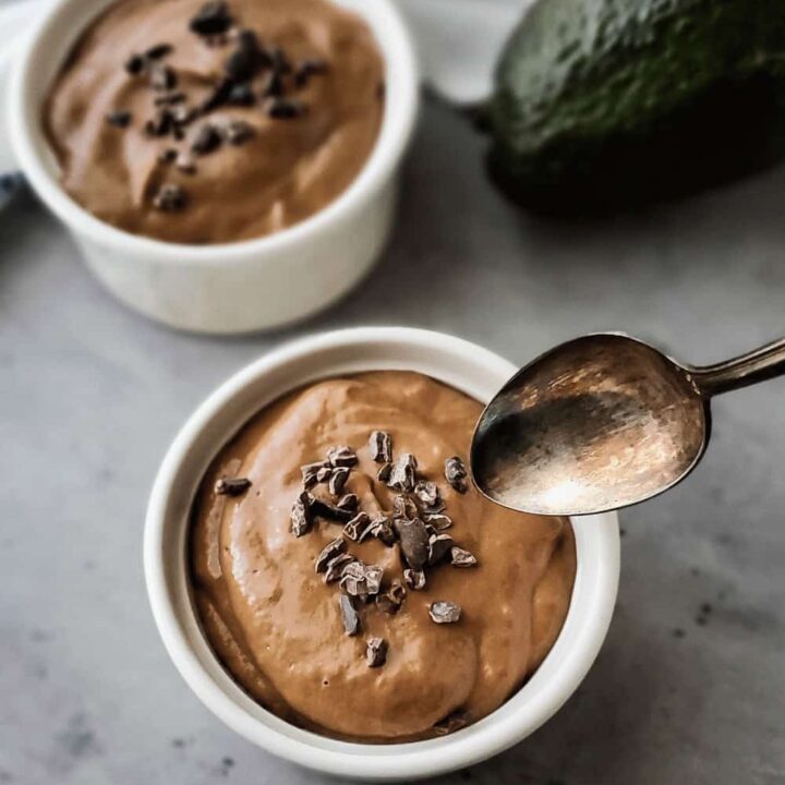 Small white bowls of pudding with raw cacao nibs sprinkled on top, a spoon hovering over one of the bowls