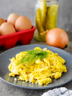 Dill pickle scrambled eggs on a gray plate garnished with fresh dill. Whole eggs, pickles, and onion in background