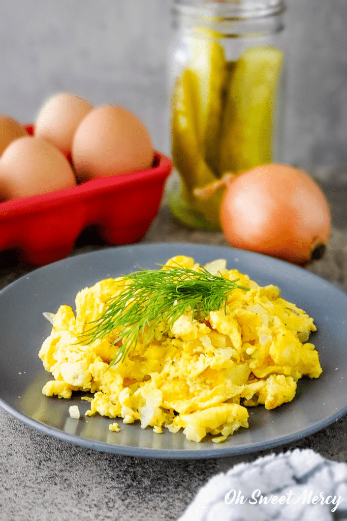 Dill pickle scrambled eggs on a gray plate garnished with fresh dill. Whole eggs, pickles, and onion in background